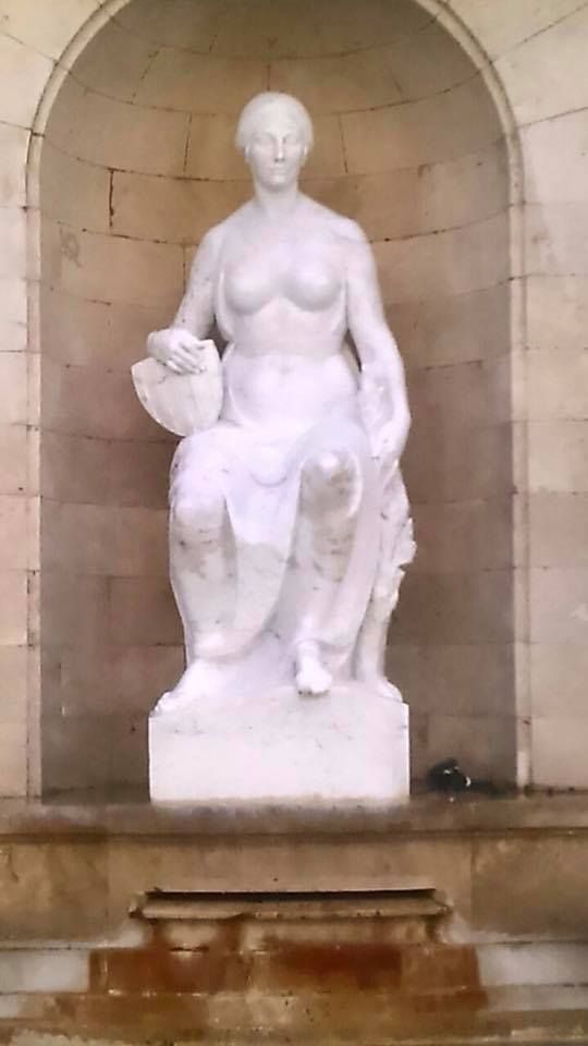 Escultura de mármol blanco que representa una figura sentada sosteniendo un objeto, situada en una alcoba arqueada de piedra.