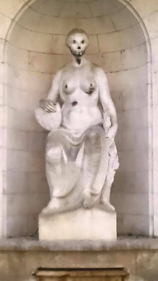 Una escultura de mármol de una figura sentada con cabeza de calavera y manchas oscuras, situada en una alcoba arqueada de piedra.