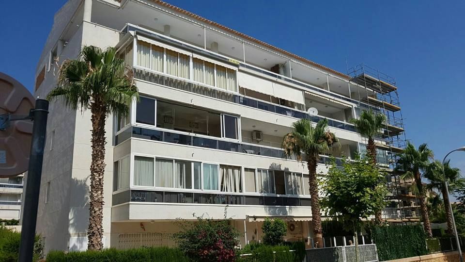 Un edificio de apartamentos blanco de varias plantas con balcones y palmeras contra un cielo azul despejado.