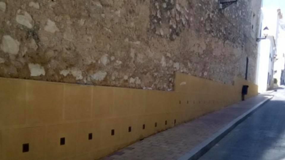 Un muro de piedra con una base pintada de amarillo que presenta una hilera de pequeños orificios de drenaje cuadrados a lo largo de una estrecha calle pavimentada.