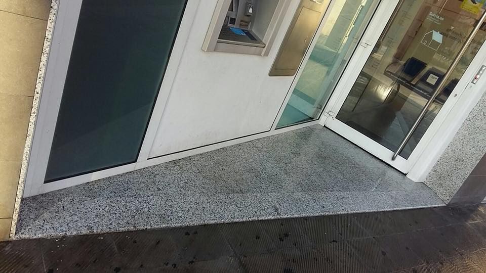 Una rampa empedrada conduce a una puerta de entrada de cristal junto a un cajero automático en una pared exterior de color claro.