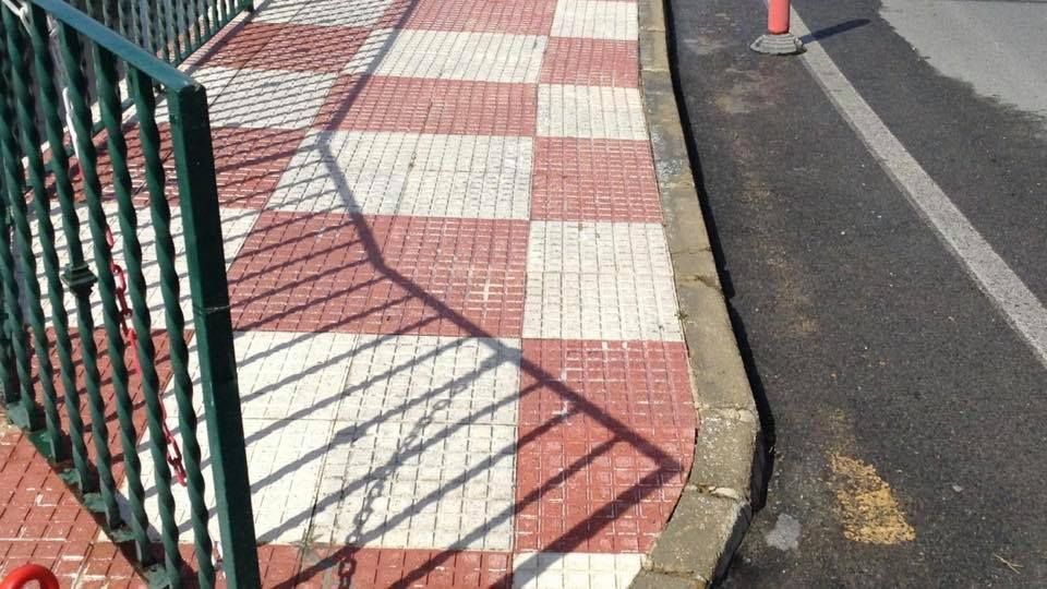 Una acera con un patrón de baldosas a cuadros rojas y blancas, junto a una valla metálica verde y una carretera pavimentada con bordillo.