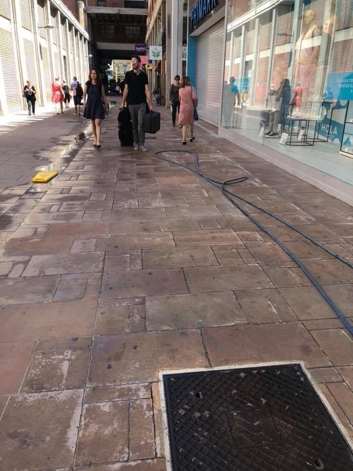 Los peatones caminan por una soleada acera urbana bordeada de escaparates y un cable de servicios públicos que cruza el sendero de piedra.