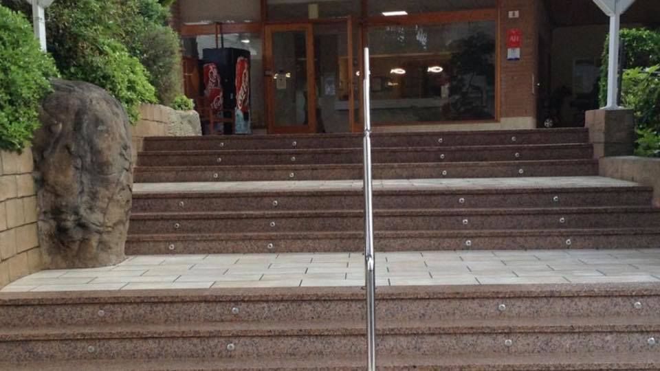 Una amplia escalera de piedra oscura conduce a una entrada con puertas de cristal y una barandilla metálica central.