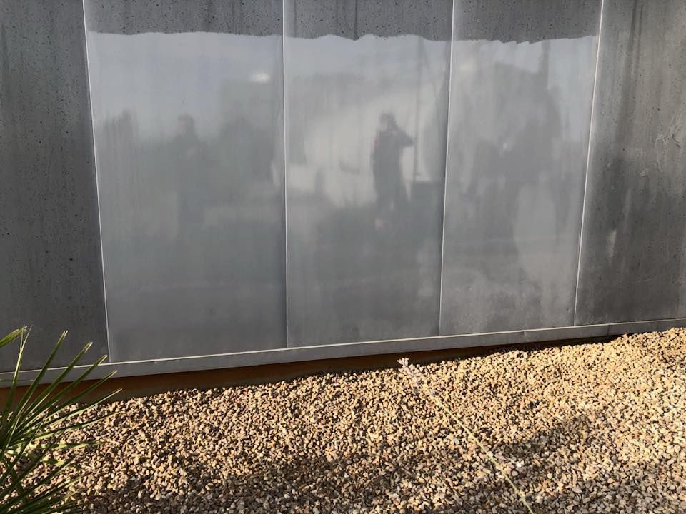 Una cortina blanca y transparente cuelga sobre una ventana, reflejando a una persona que se encuentra afuera contra un fondo de grava.