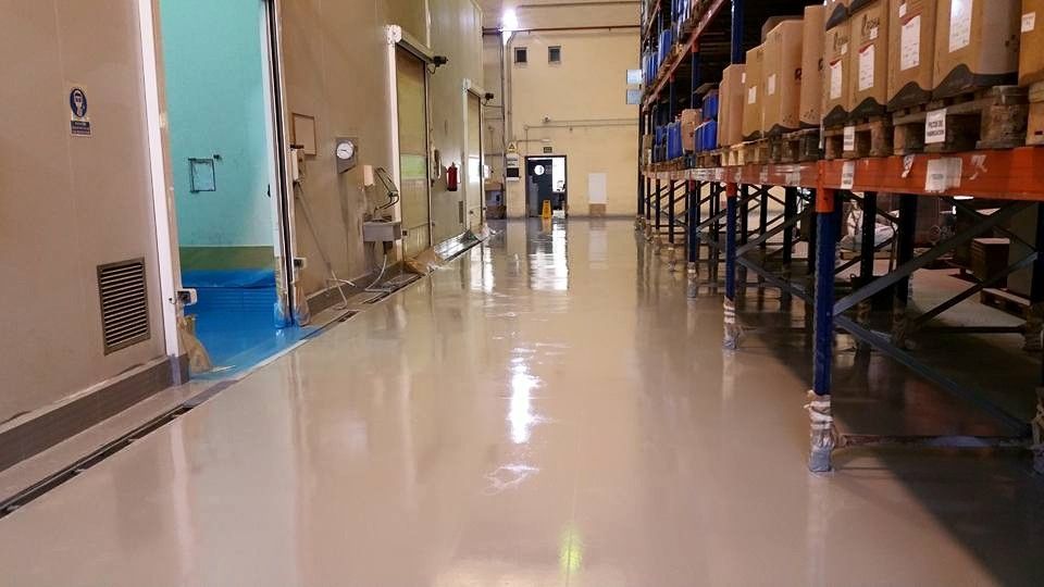 Vista de un pasillo de un almacén con suelo de resina epoxi gris claro brillante y estanterías metálicas a la derecha.