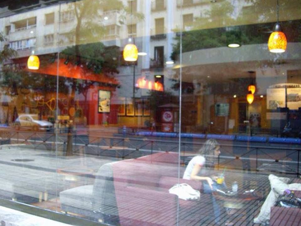 Una persona está sentada en una mesa de una cafetería dentro de un edificio con fachada de cristal, con una calle de la ciudad reflejada en la ventana.