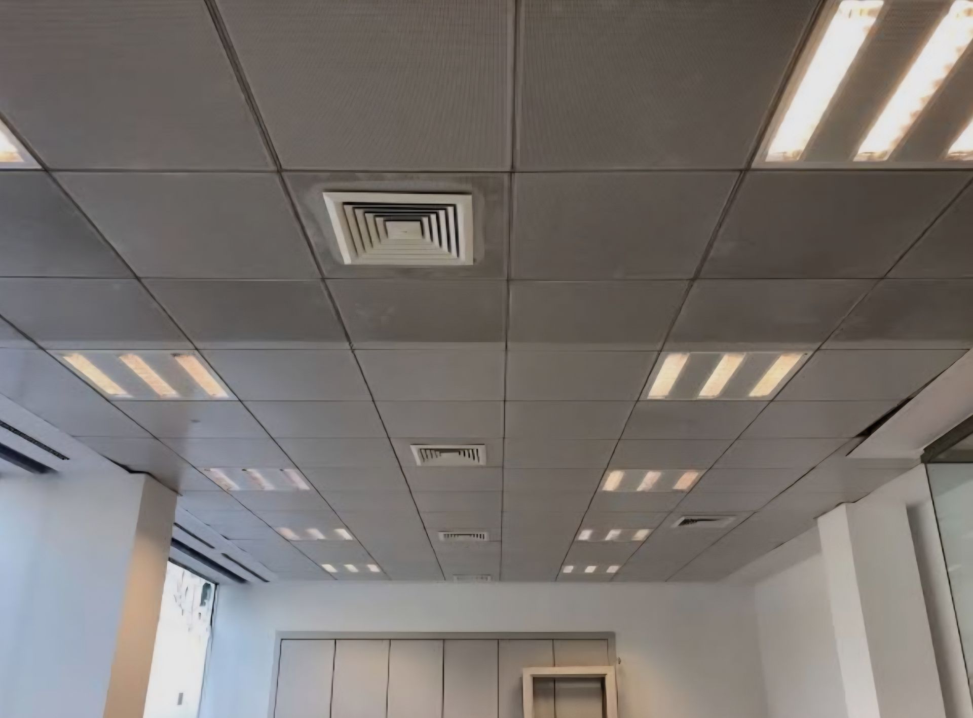 Vista de un techo de rejilla gris con paneles de iluminación empotrados y rejillas de ventilación cuadradas.