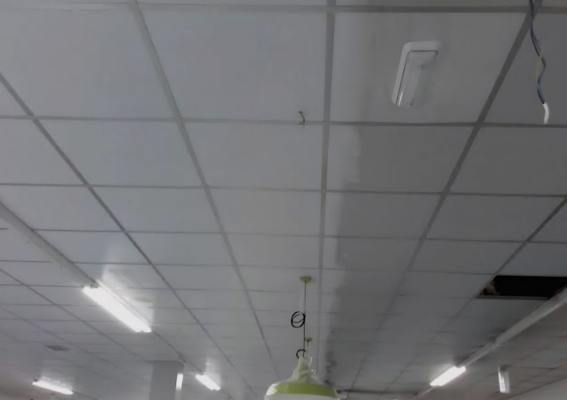 Un techo suspendido blanco con baldosas cuadradas, luminarias de techo, una lámpara colgante y un panel de techo faltante.