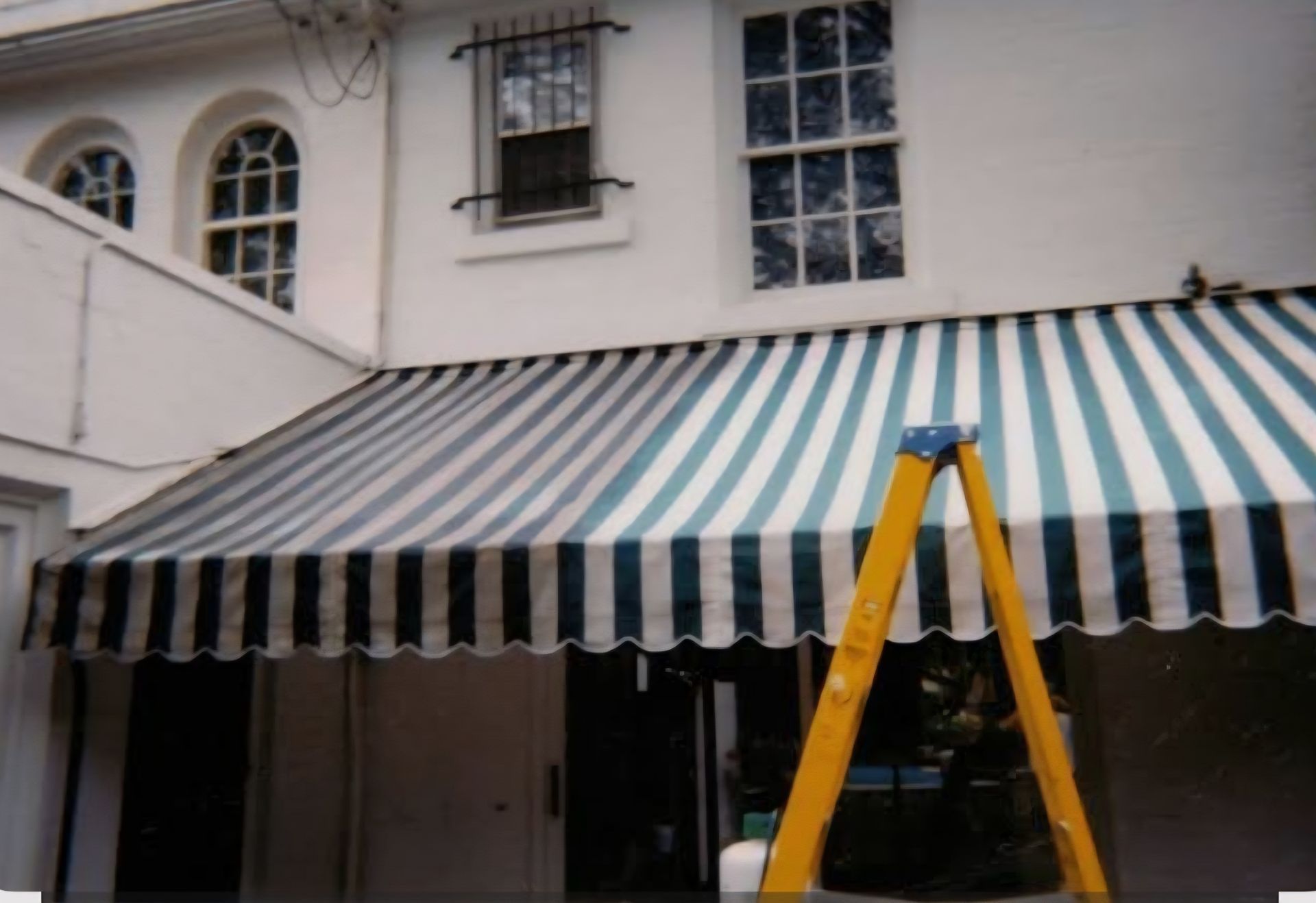 Una escalera amarilla se alza frente a la pared blanca de un edificio con ventanas y un toldo a rayas con bordes festoneados.