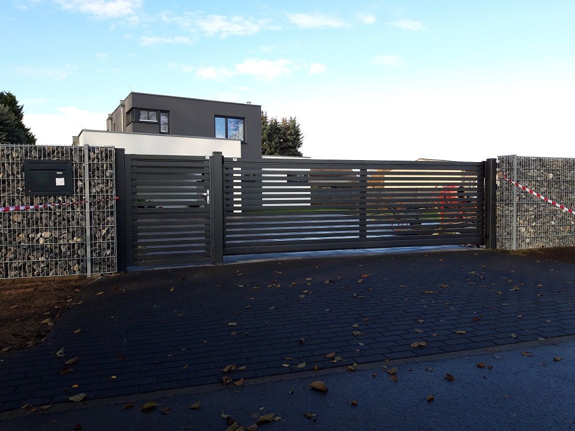 Graues modernes Tor und Zaun mit einer Gabionenwand neben einem Haus, schwarz gepflasterte Auffahrt.