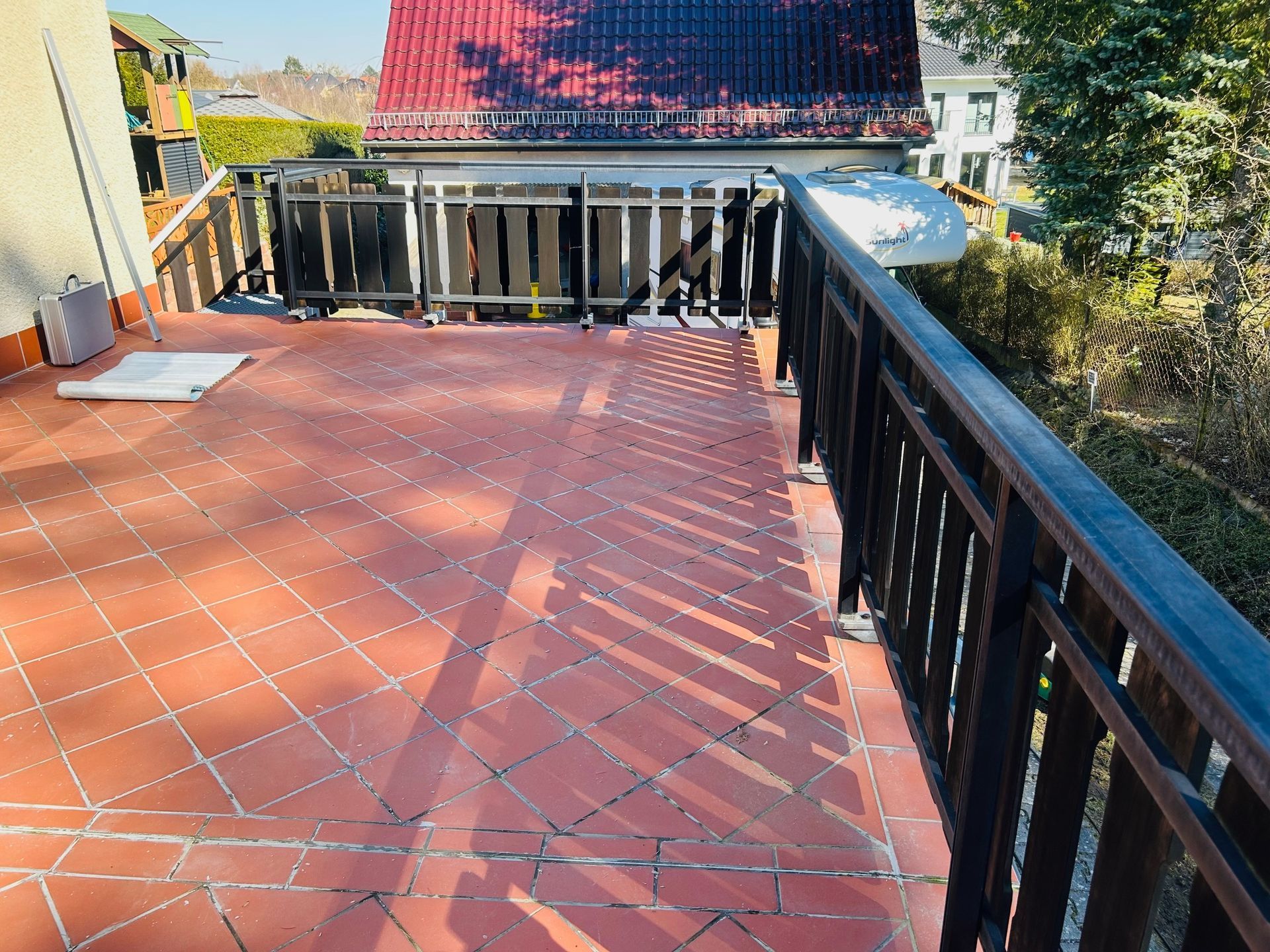 Eine sonnige Backsteinterrasse mit dunkelbraunem Geländer. Im Hintergrund ist ein Haus mit rotem Dach zu sehen.