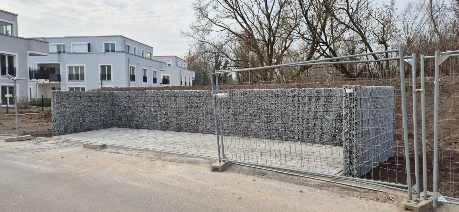 Eine Baustelle mit einer Gabionenwand und einem provisorischen Zaun. Im Hintergrund sind moderne weiße Gebäude zu sehen.