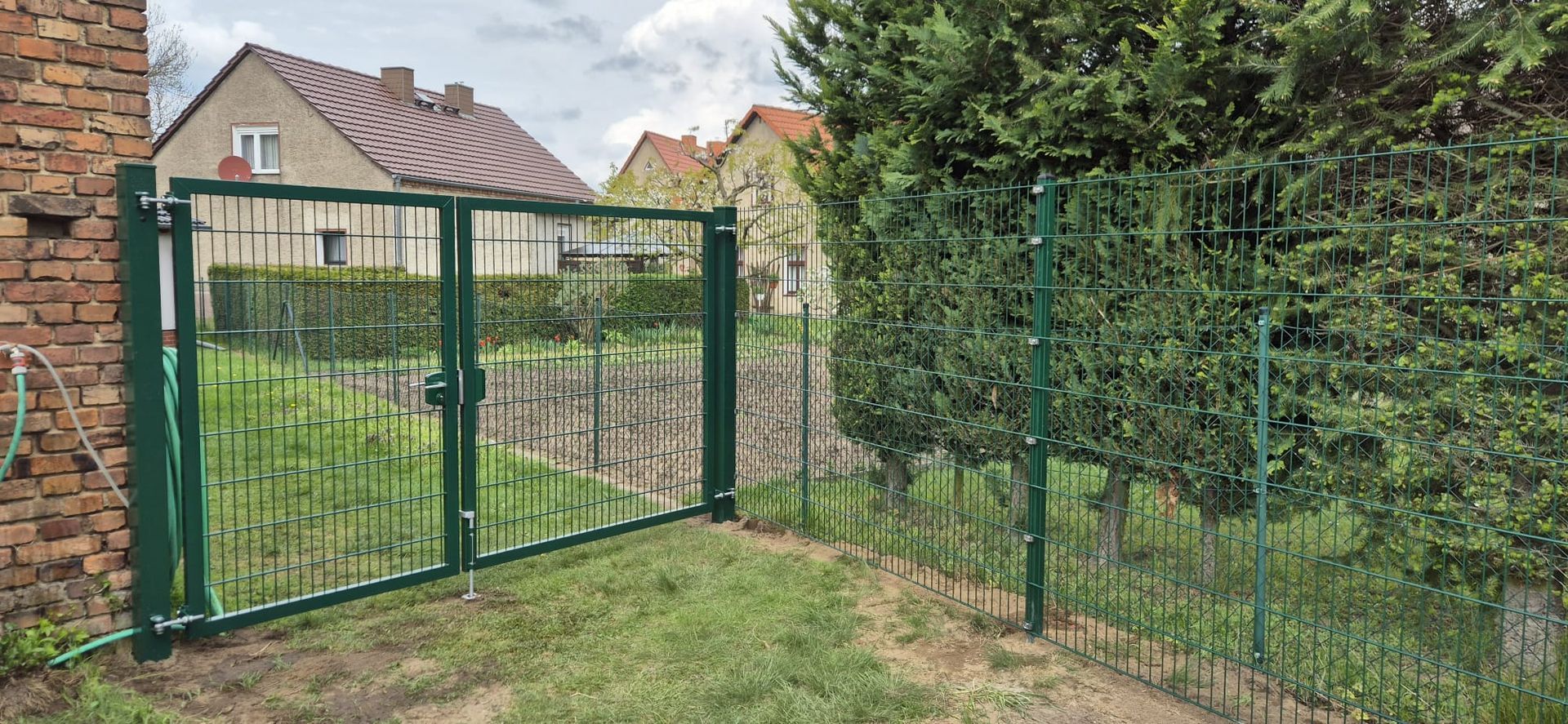 Grünes Metalltor in einem grasbewachsenen Hof mit Backsteinmauer links und einem grünen Zaun und Büschen rechts. Im Hintergrund sind Häuser zu sehen.