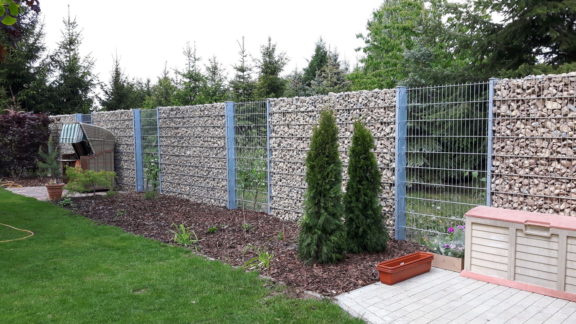 Ein mit Steinen gefüllter Gabionenzaun in einem Hinterhof, flankiert von Bäumen, Grünflächen und einem kleinen Garten.