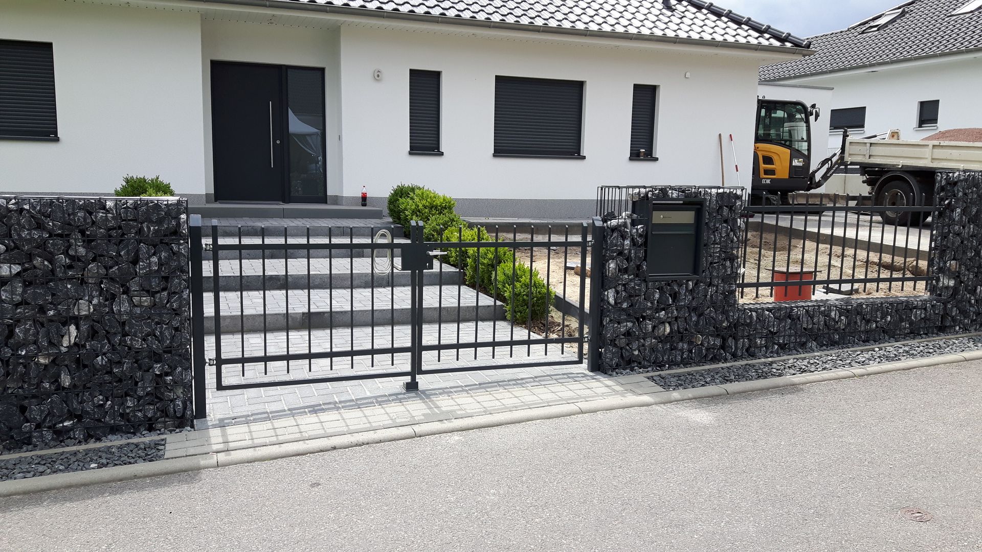 Ein modernes Haus mit schwarzem Metallzaun und Steinmauern. Im Hintergrund ist ein kleiner Bagger zu sehen.