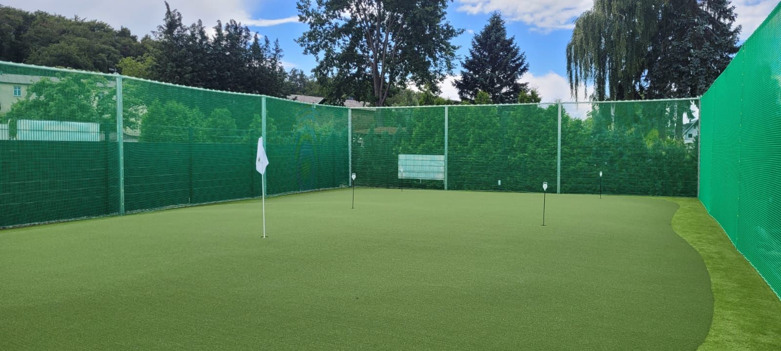 Ein Spielfeld, umgeben von einem grünen Netzzaun und Bäumen. Weiße Fahnen markieren die Löcher auf dem Grün, über dem sich ein blauer Himmel befindet.