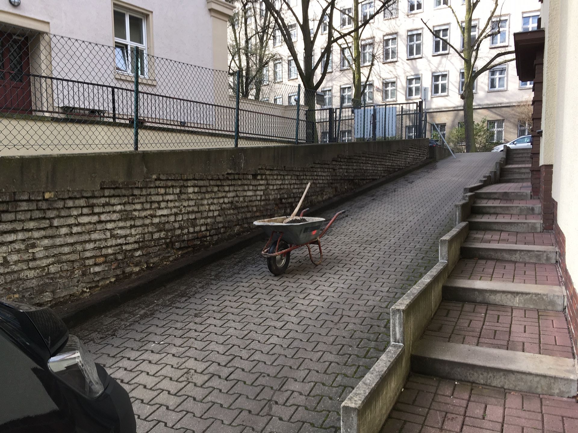 Schubkarre auf einer nach oben führenden Ziegelrampe neben der Treppe. Links eine Ziegelmauer, im Hintergrund ein Gebäude.