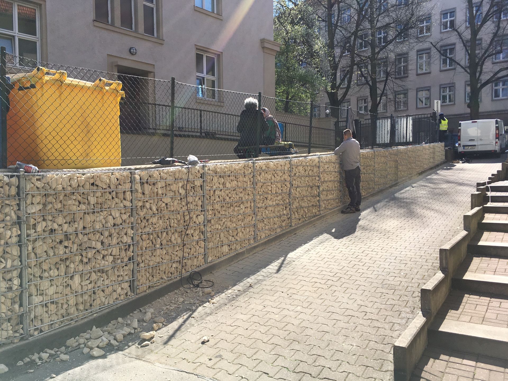 Arbeiter errichten eine Steinmauer entlang eines Gehwegs. Die Mauer besteht aus Drahtkäfigen, die mit hellen Steinen gefüllt sind. Daneben steht ein gelber Bausack.