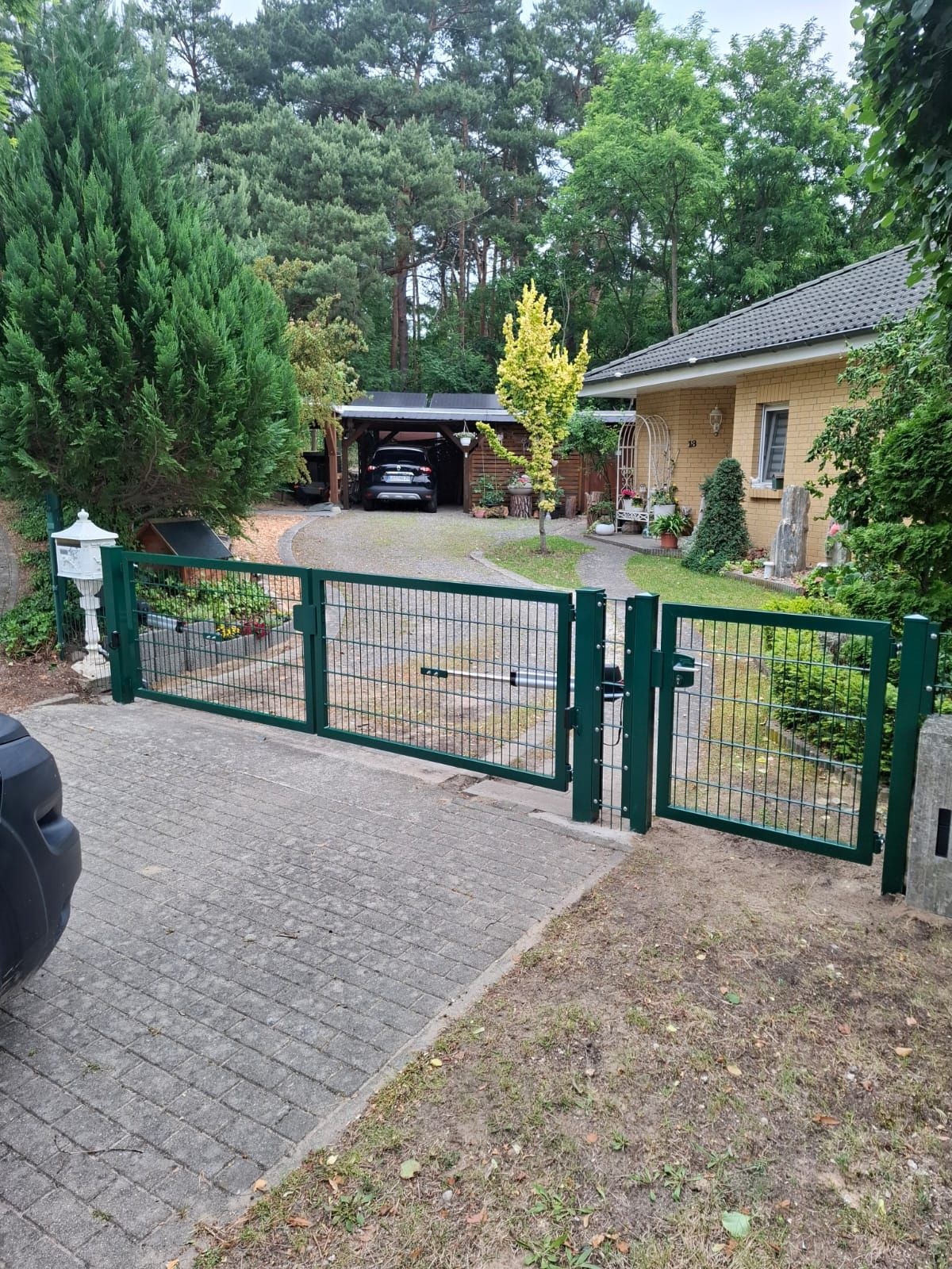 Grünes Tor und Zaun vor einem Haus mit Auffahrt, Briefkasten und Garage. Bäume umgeben das Grundstück.