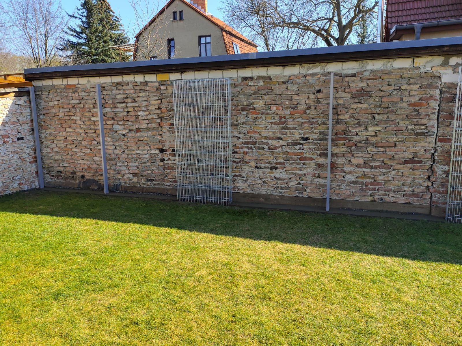 Eine Ziegelmauer mit Metallstützen in einem grasbewachsenen Hof. Ein Bündel Metallstangen lehnt an der Mauer.