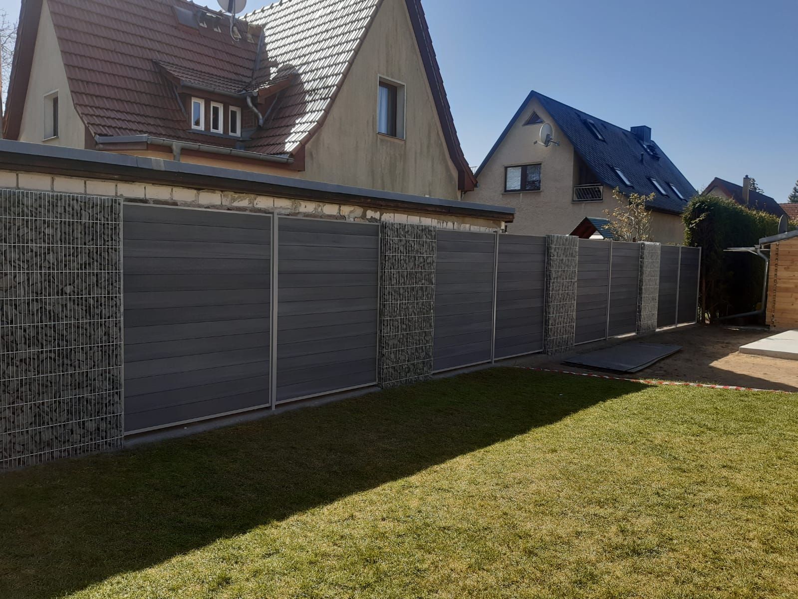 Ein grauer Sichtschutzzaun mit Steinsäulen in einem Hinterhof. Der Zaun trennt einen Rasen von Häusern mit Ziegeldächern.