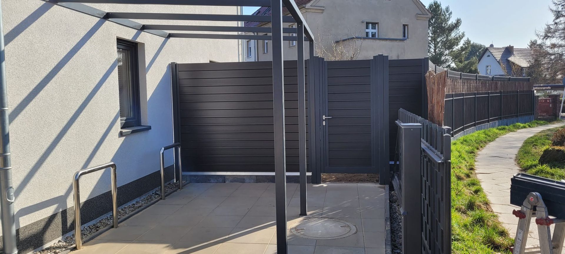 Ein eingezäunter Terrassenbereich mit Betonboden, einem schwarzen Tor und einem Gehweg, der an einem sonnigen Tag zu einem Haus führt.