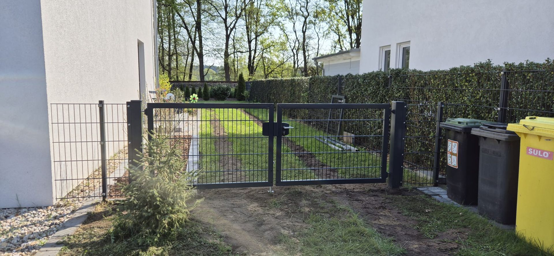 Schwarzes Metalltor, das in einen Hof führt, mit einer Hecke an der Rückseite, einem weißen Haus auf beiden Seiten und Mülltonnen auf der rechten Seite.