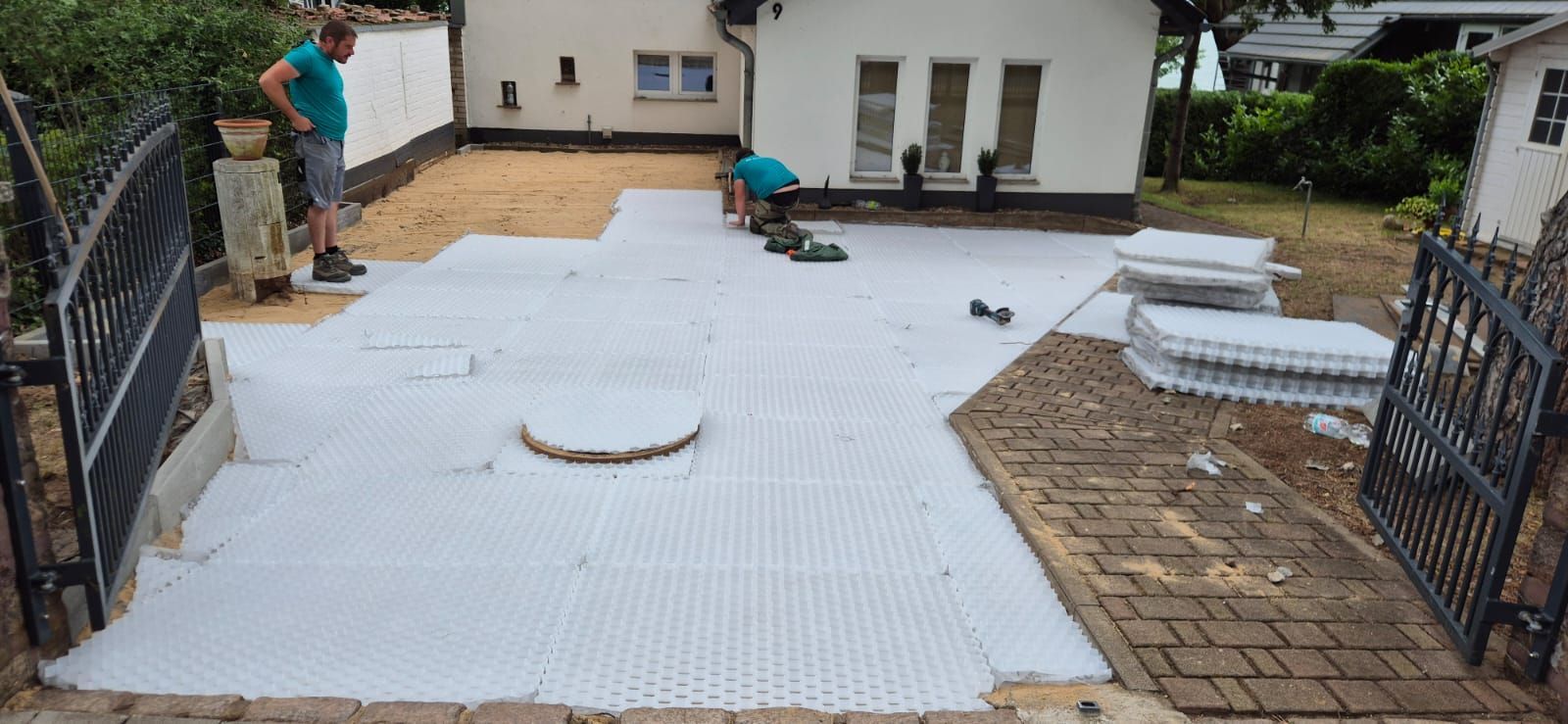 Zwei Männer arbeiten auf einer Terrasse. Weiße Plastikgitter sind ausgelegt, Mauerwerk und ein Haus sind zu sehen.