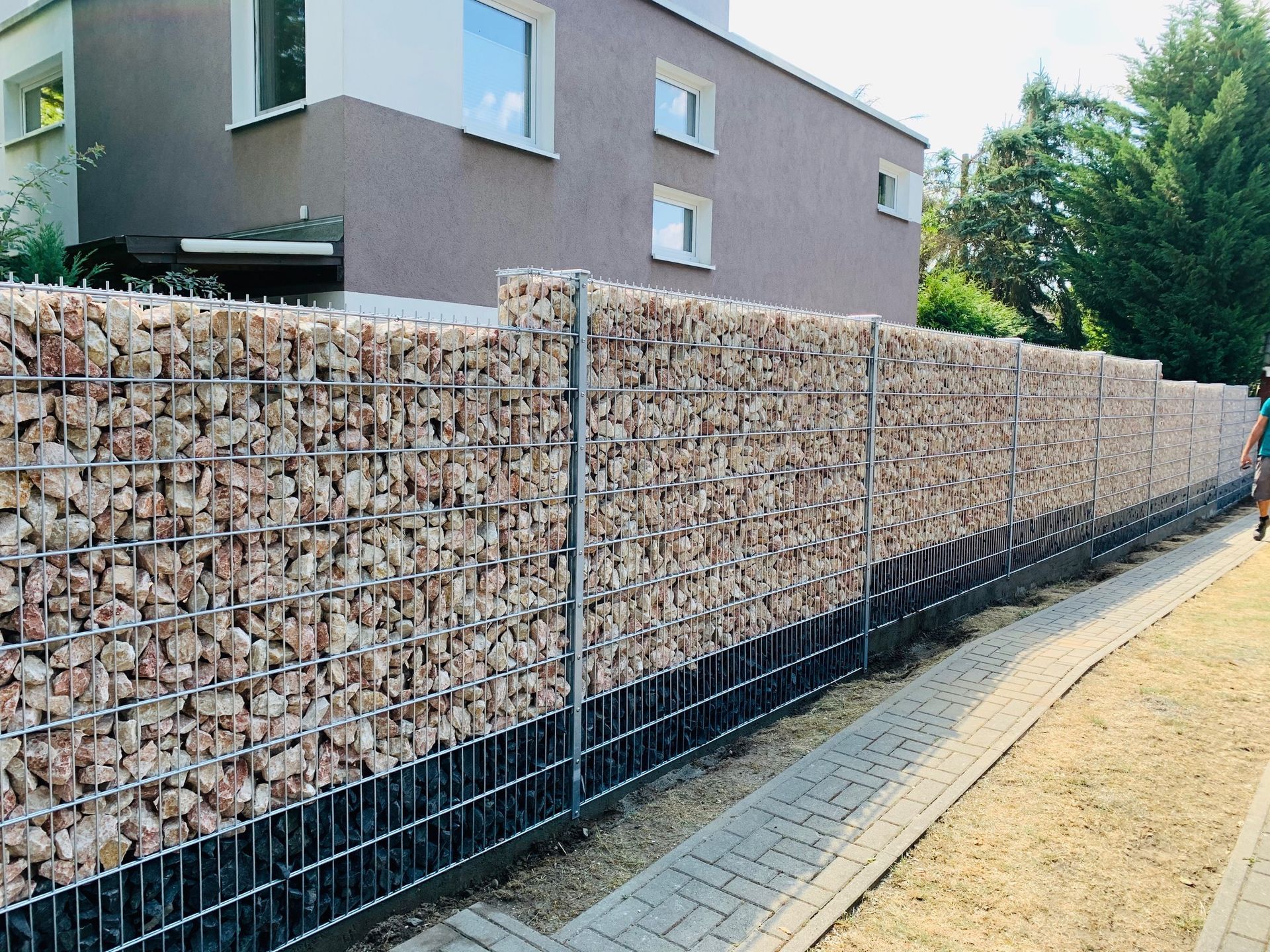 Ein langer Gabionenzaun mit hellbraunen Steinen säumt einen Bürgersteig und grenzt an ein Haus mit brauner Fassade.