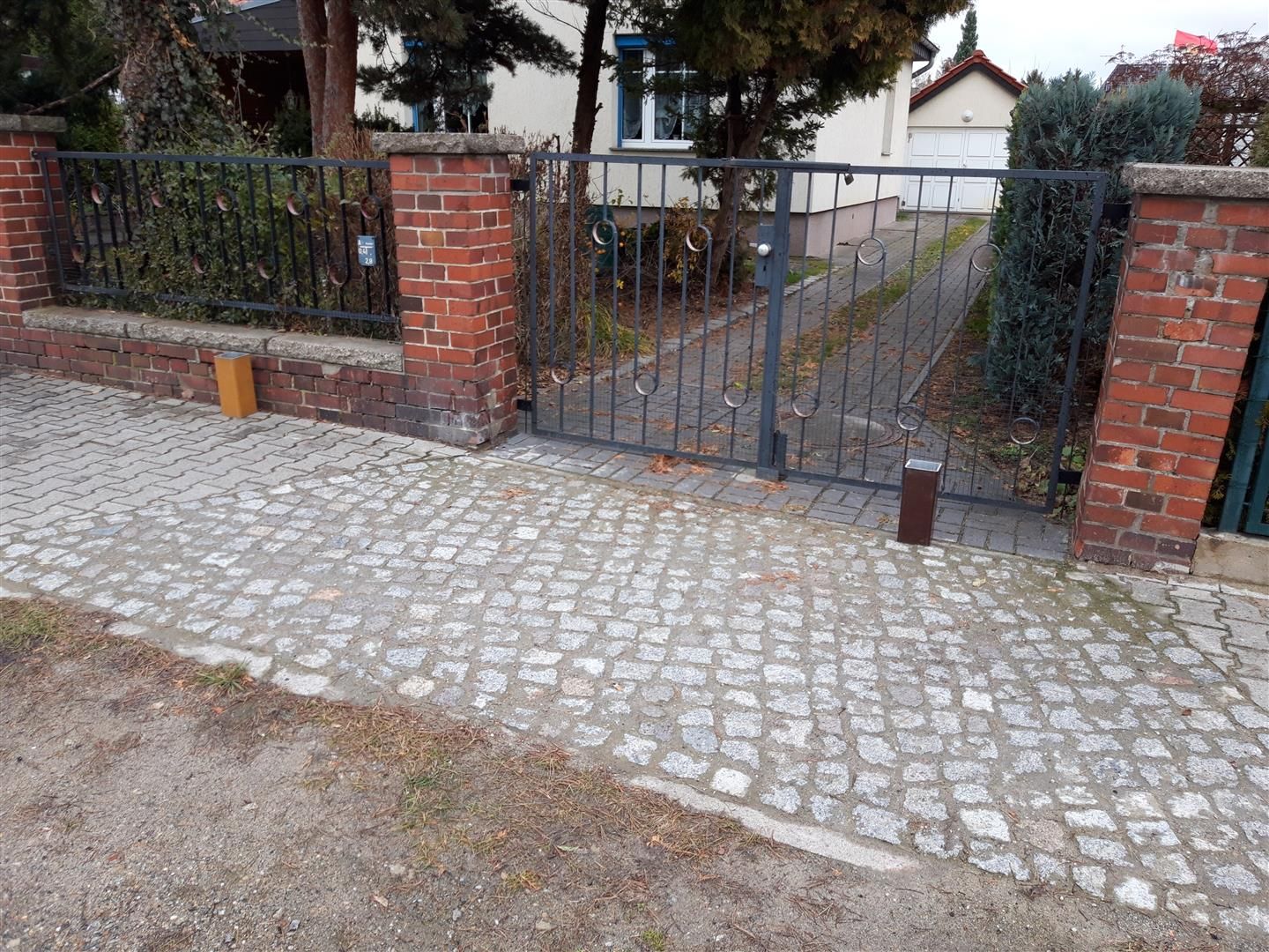 Ziegelpfeiler rahmen ein schwarzes Metalltor ein, das zu einer gepflasterten Auffahrt führt. Im Hintergrund sind ein Haus und Grünflächen zu sehen.