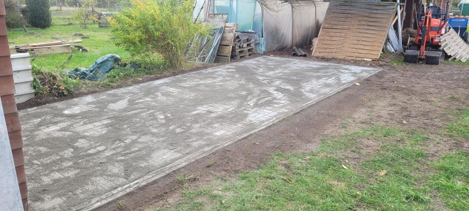 Eine Betonplatte in einem Hof mit einem Schuppen und einem Bagger im Hintergrund. Grünes Gras umgibt den Beton.