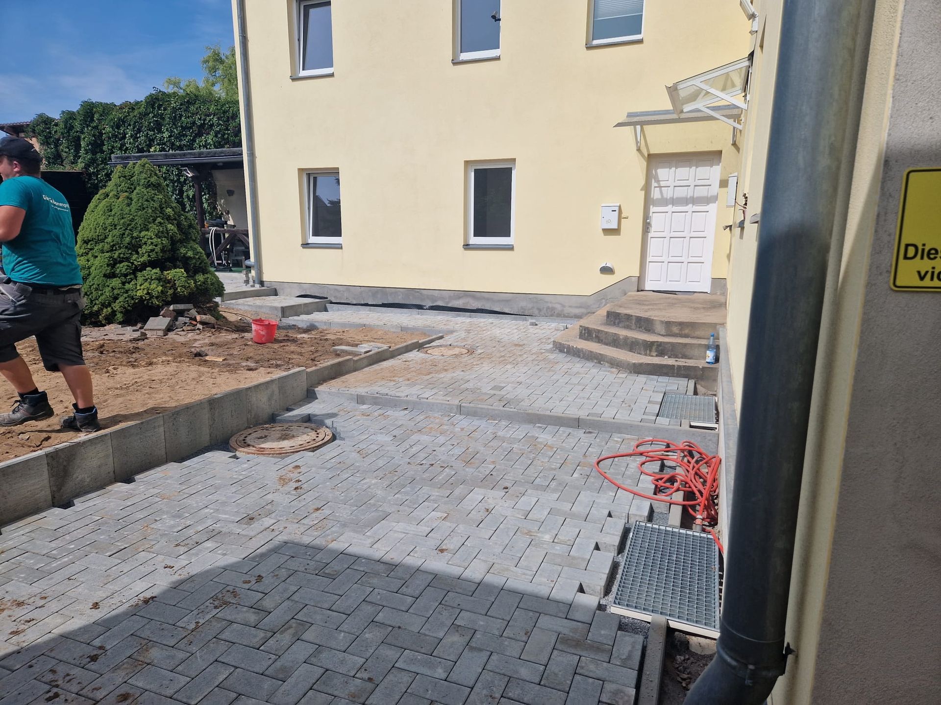 Bauarbeiter pflastert einen Hof vor einem gelben Haus. Graue Pflastersteine, Erde und eine kleine Stützmauer sind zu sehen.