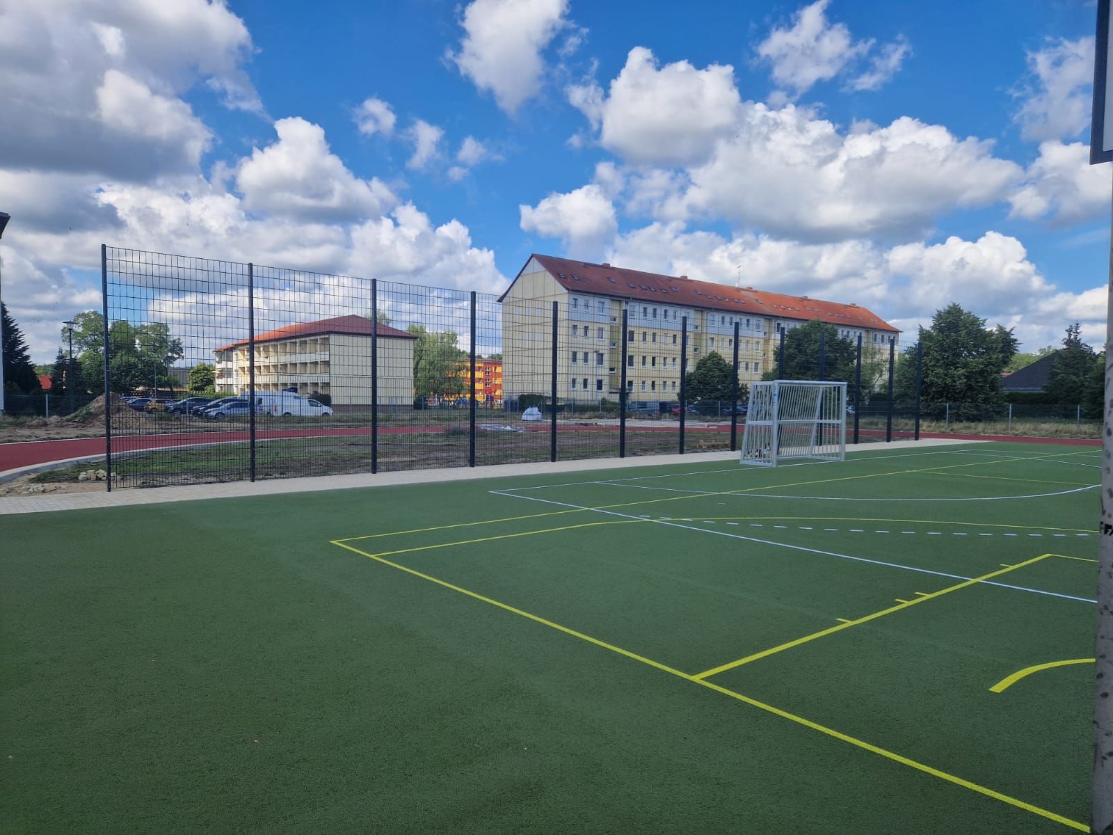Ein grüner Sportplatz mit gelben Linien vor einem mehrstöckigen Gebäude unter einem blauen Himmel mit flauschigen weißen Wolken.