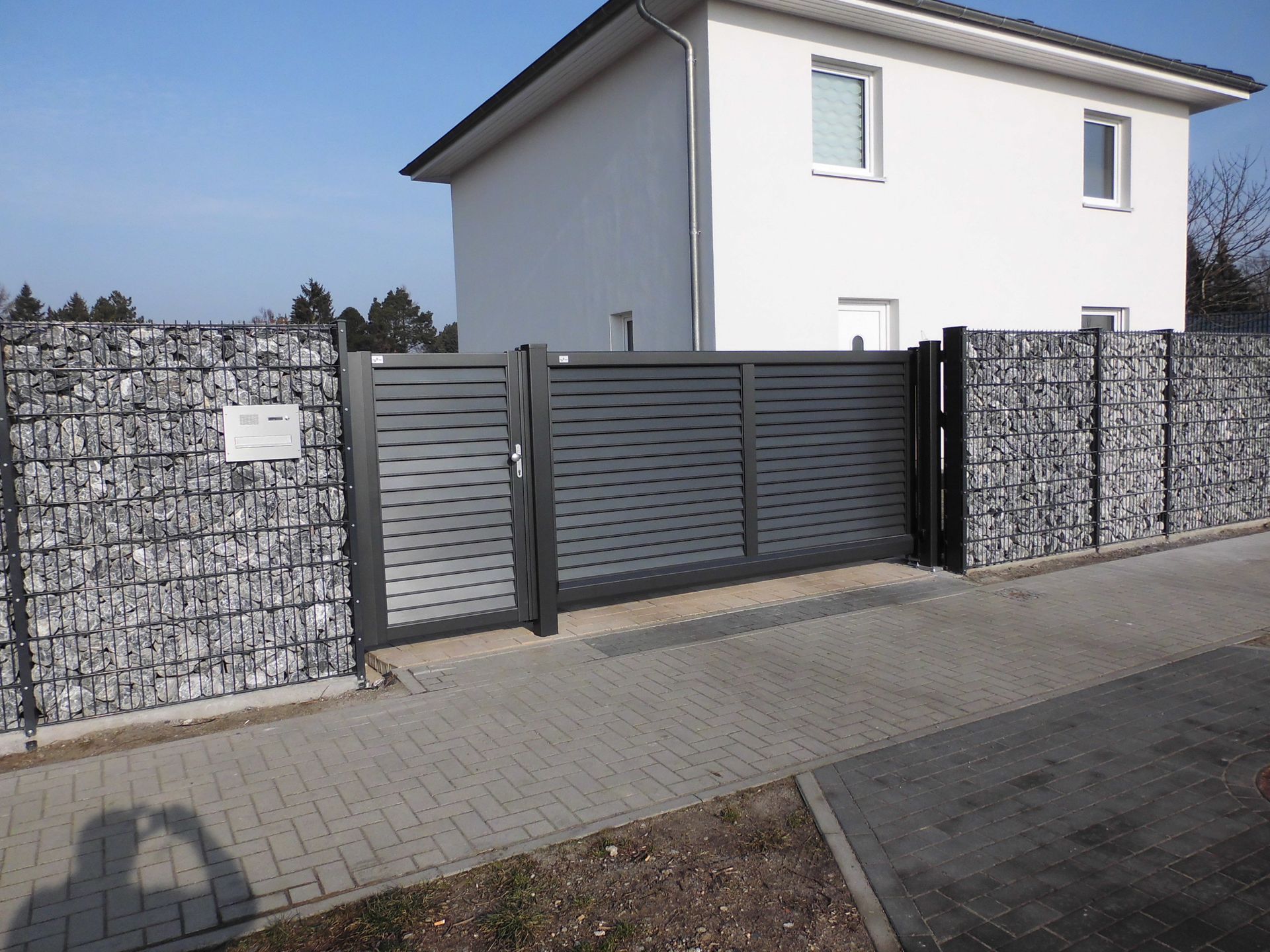 Ein graues, modernes Tor und ein Zaun aus Stein und horizontalen Latten vor einem zweistöckigen weißen Haus.