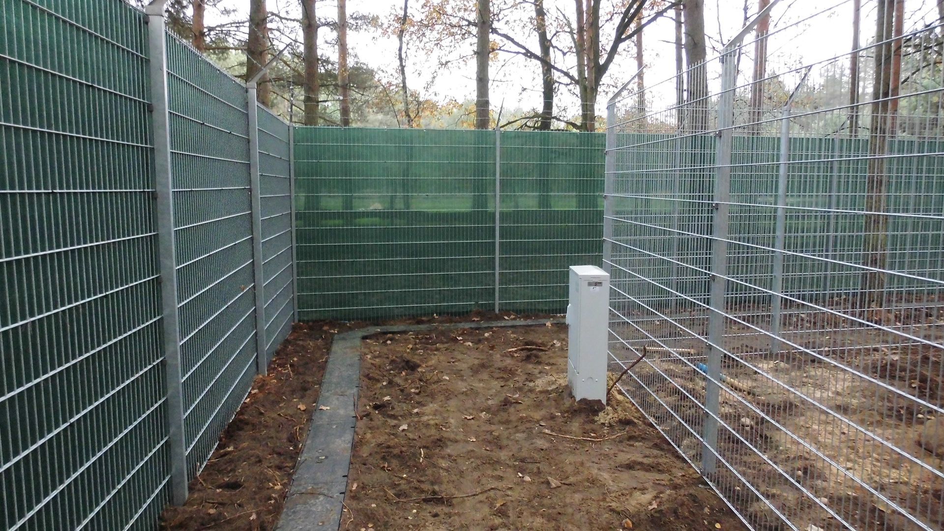 Ein rechteckiger Bereich, der durch einen Metallzaun begrenzt wird. Der Zaunabschnitt ist grün abgeschirmt und mit brauner Erde gefüllt.