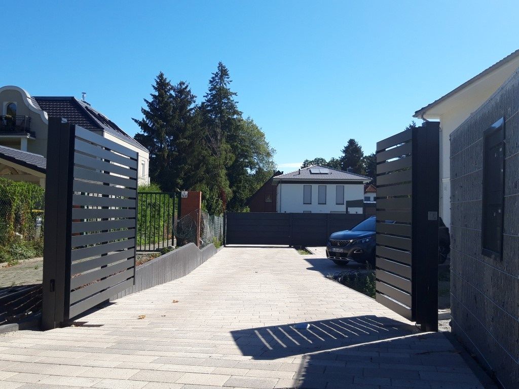 Offene, schwarze Metalltore führen zu einer Einfahrt. Ein Auto ist im Inneren geparkt, im Hintergrund sind Häuser und Bäume zu sehen.