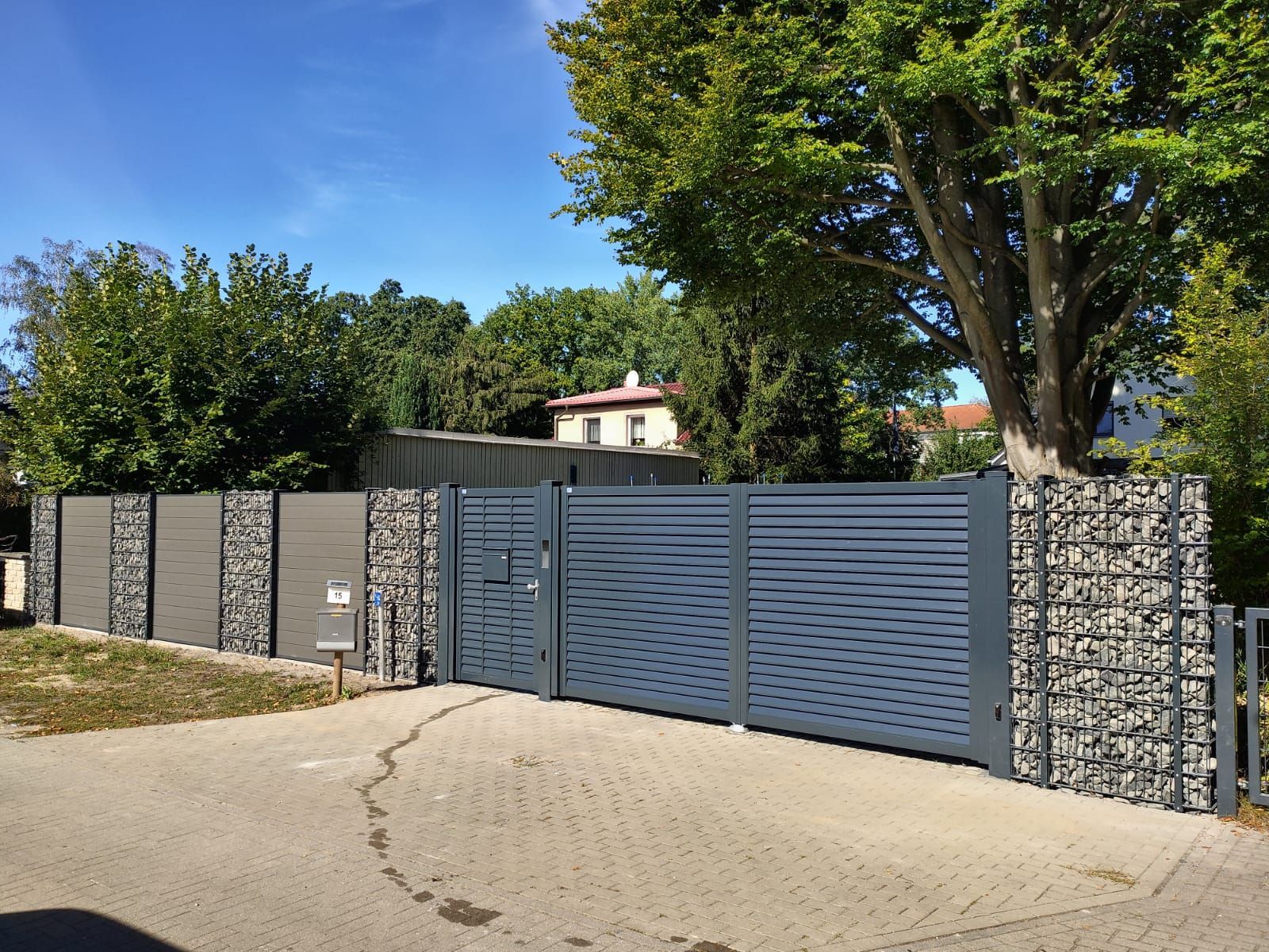 Ein modernes graues Tor und ein Zaun mit Steinakzenten umschließen eine Auffahrt. Im Hintergrund sind Bäume und ein Haus zu sehen.
