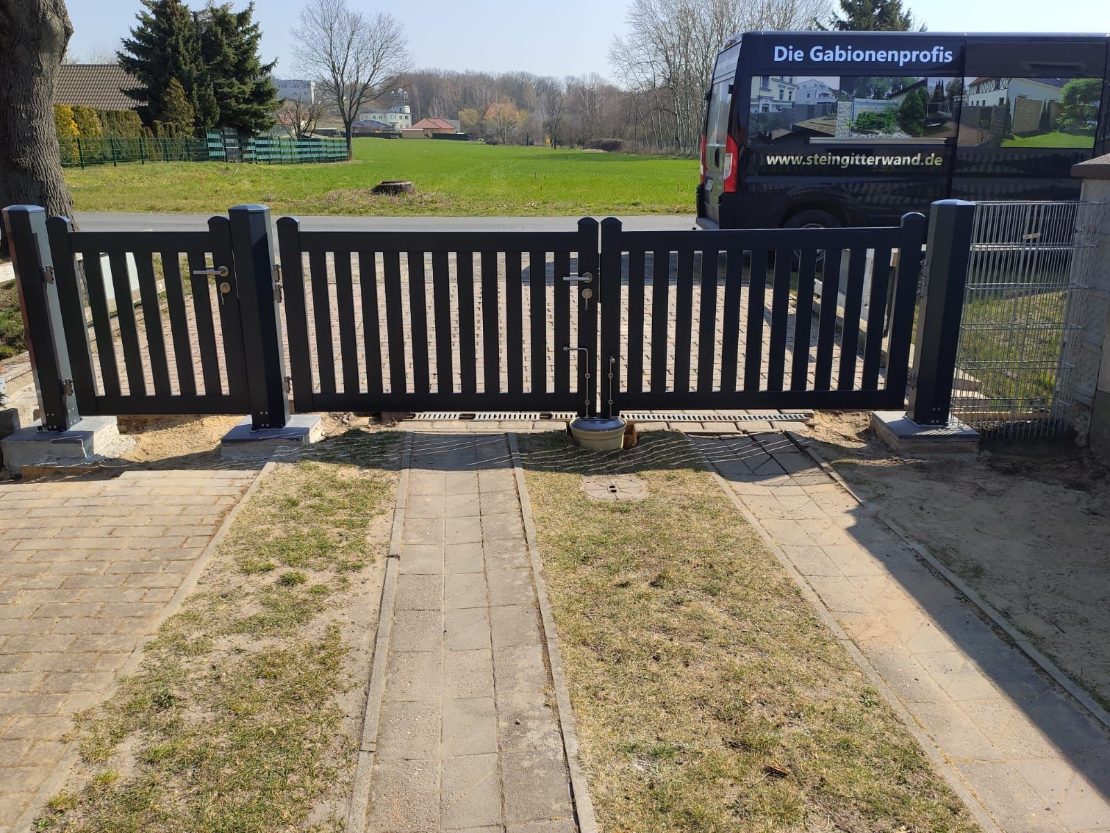 Schwarzes Holztor an einer gepflasterten Auffahrt, die zu einer Rasenfläche führt. In der Nähe parkt ein schwarzer Lieferwagen.