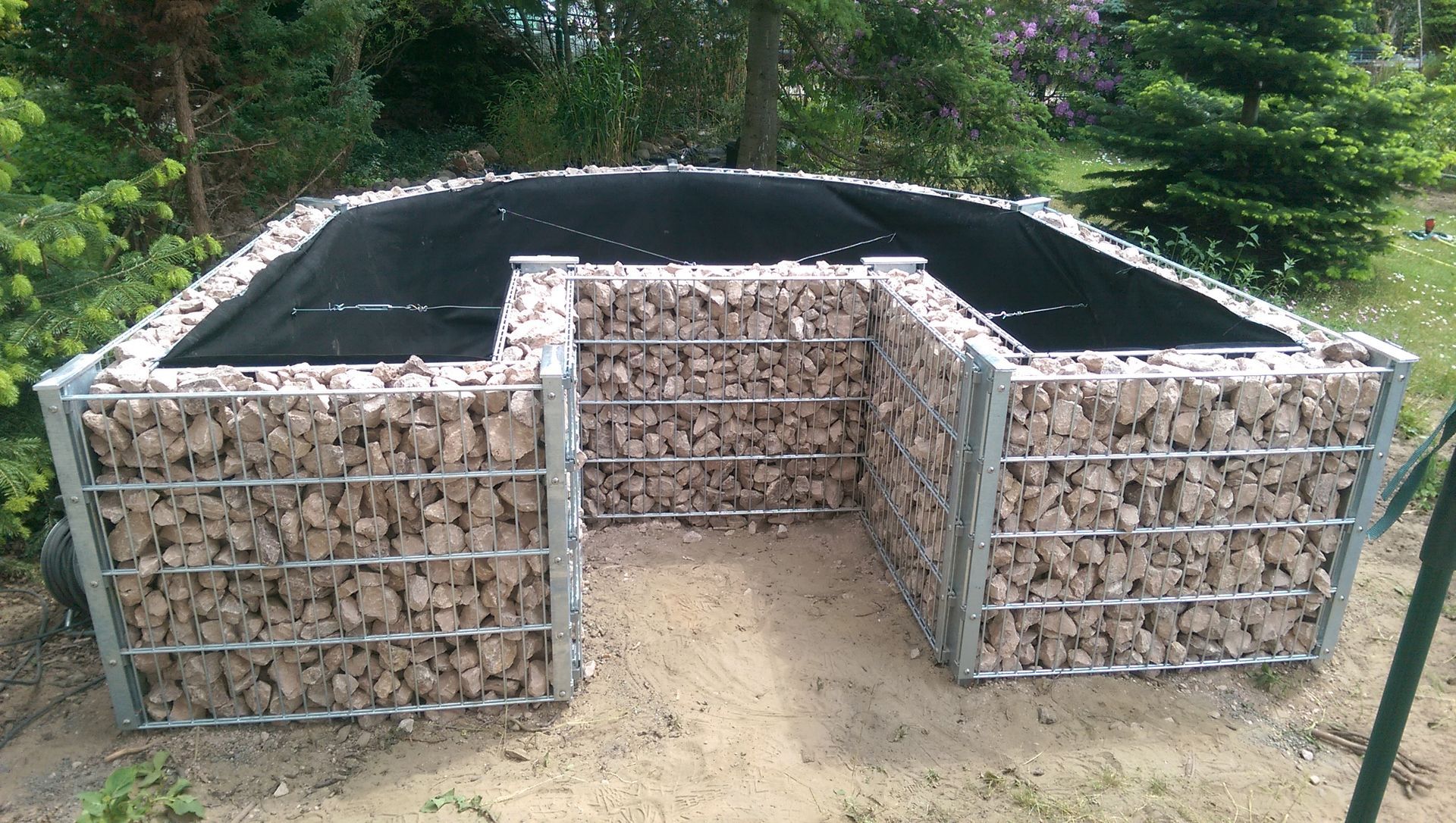 Eine U-förmige Stützmauer aus Drahtkörben, gefüllt mit hellen Steinen. Die Oberseite ist mit schwarzem Stoff bedeckt, im Freien aufgestellt.