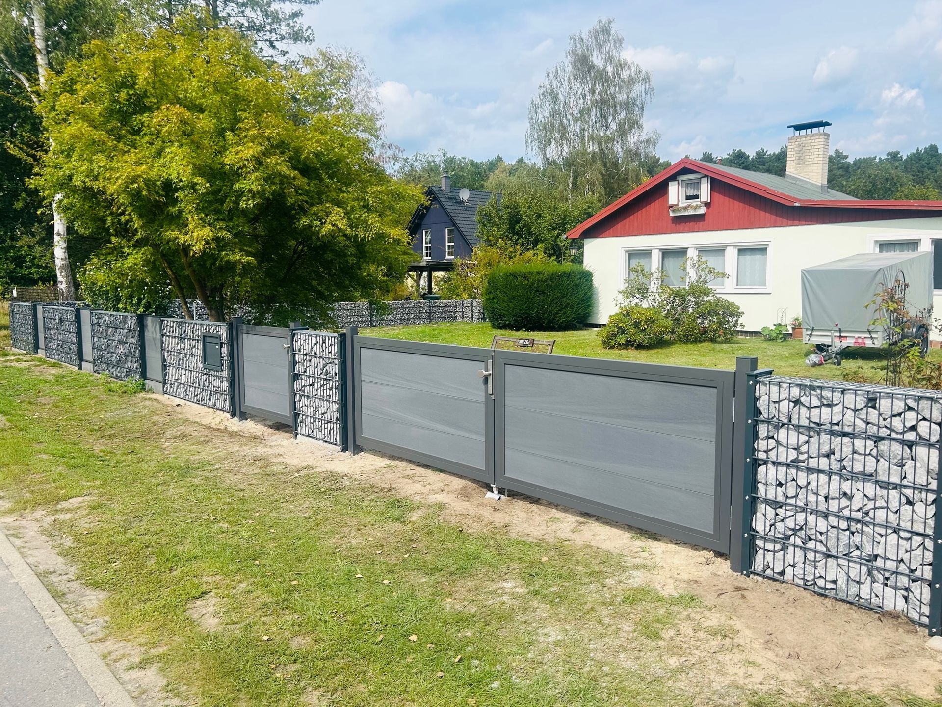 Ein grauer und steinerner Zaun umschließt einen Hof mit einem Tor vor einem Haus mit rotem Dach und einem Baum im Hintergrund.