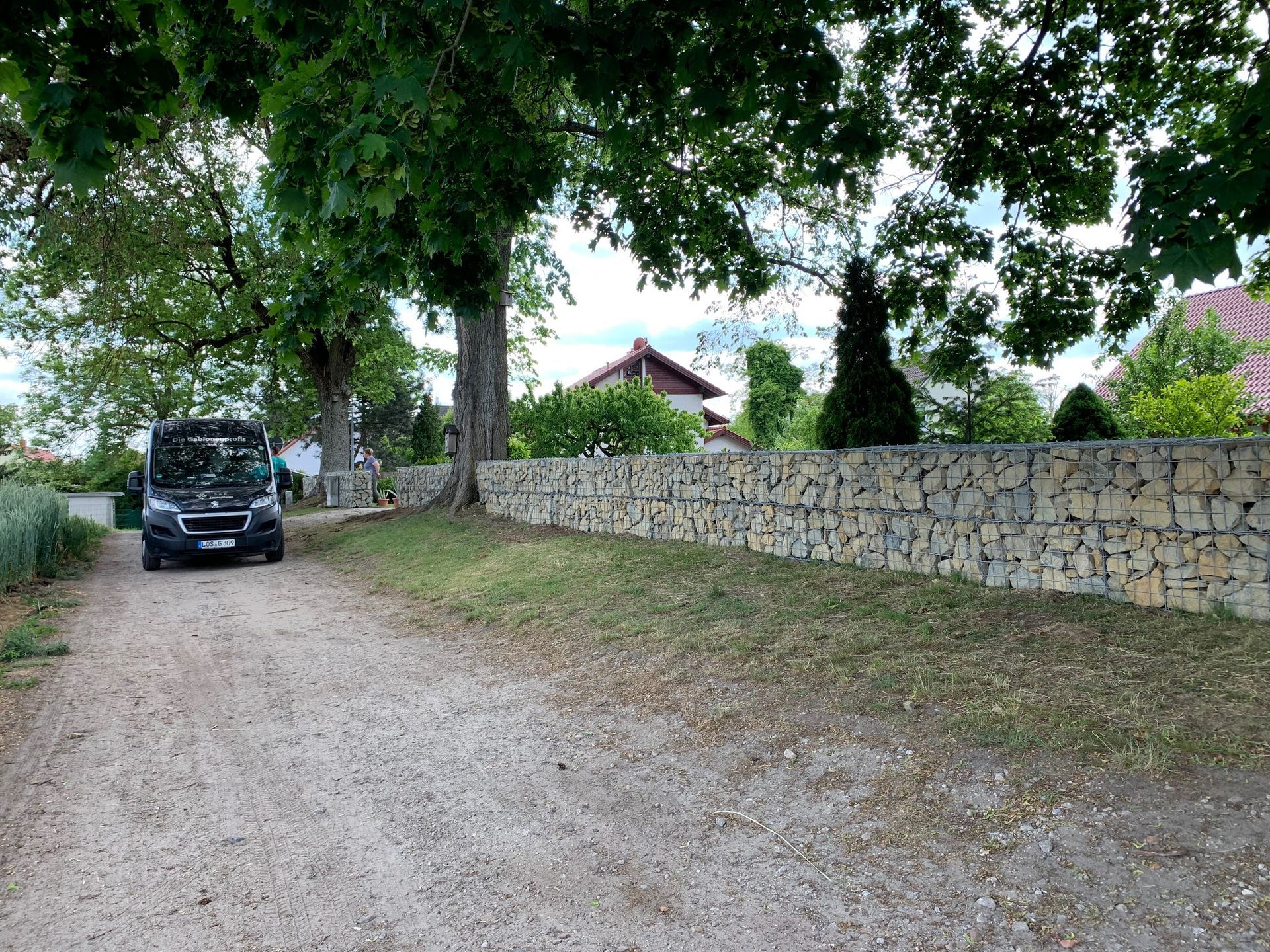 Ein schwarzer Lieferwagen parkt auf einer Schotterstraße neben einer Steinmauer unter einem großen Baum. Grünes Gras und ein Haus sind zu sehen.