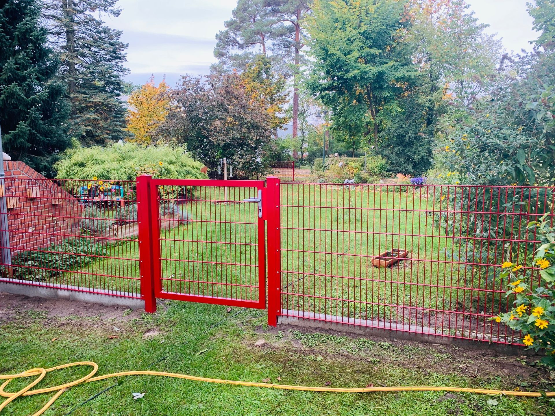 Rotes Tor und Zaun in einem grünen Hof mit Bäumen und einem bewölkten Himmel im Hintergrund.