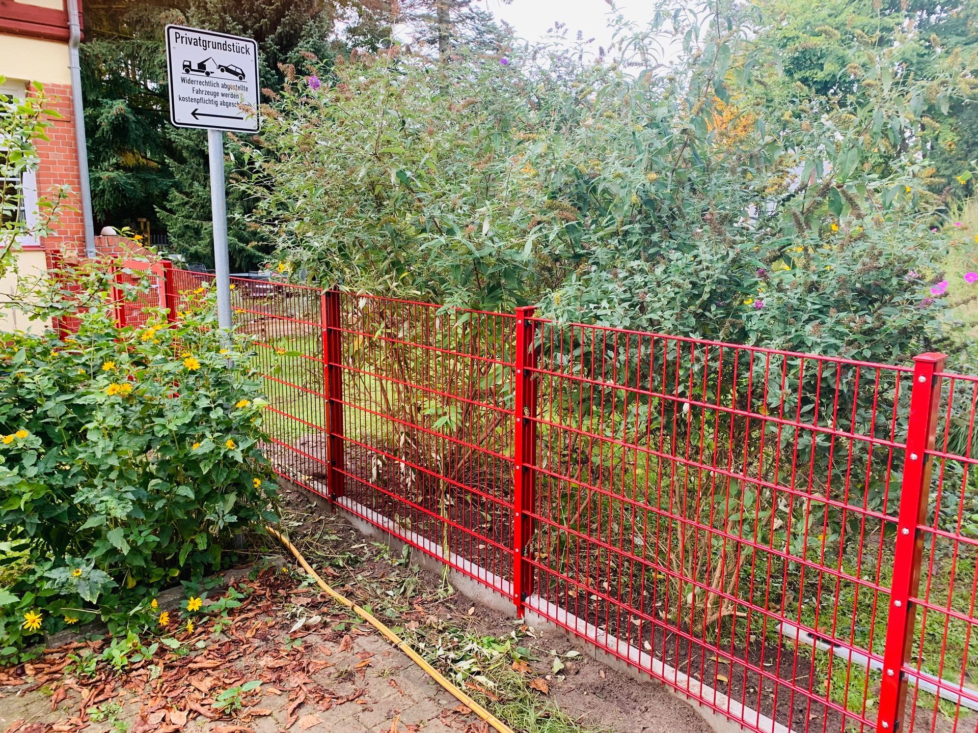 Roter Drahtzaun begrenzt einen Garten mit einem Wegweiser und üppigem grünem Laub. Der Zaun hat einen roten Rahmen und ein dunkelrotes Drahtgeflecht.