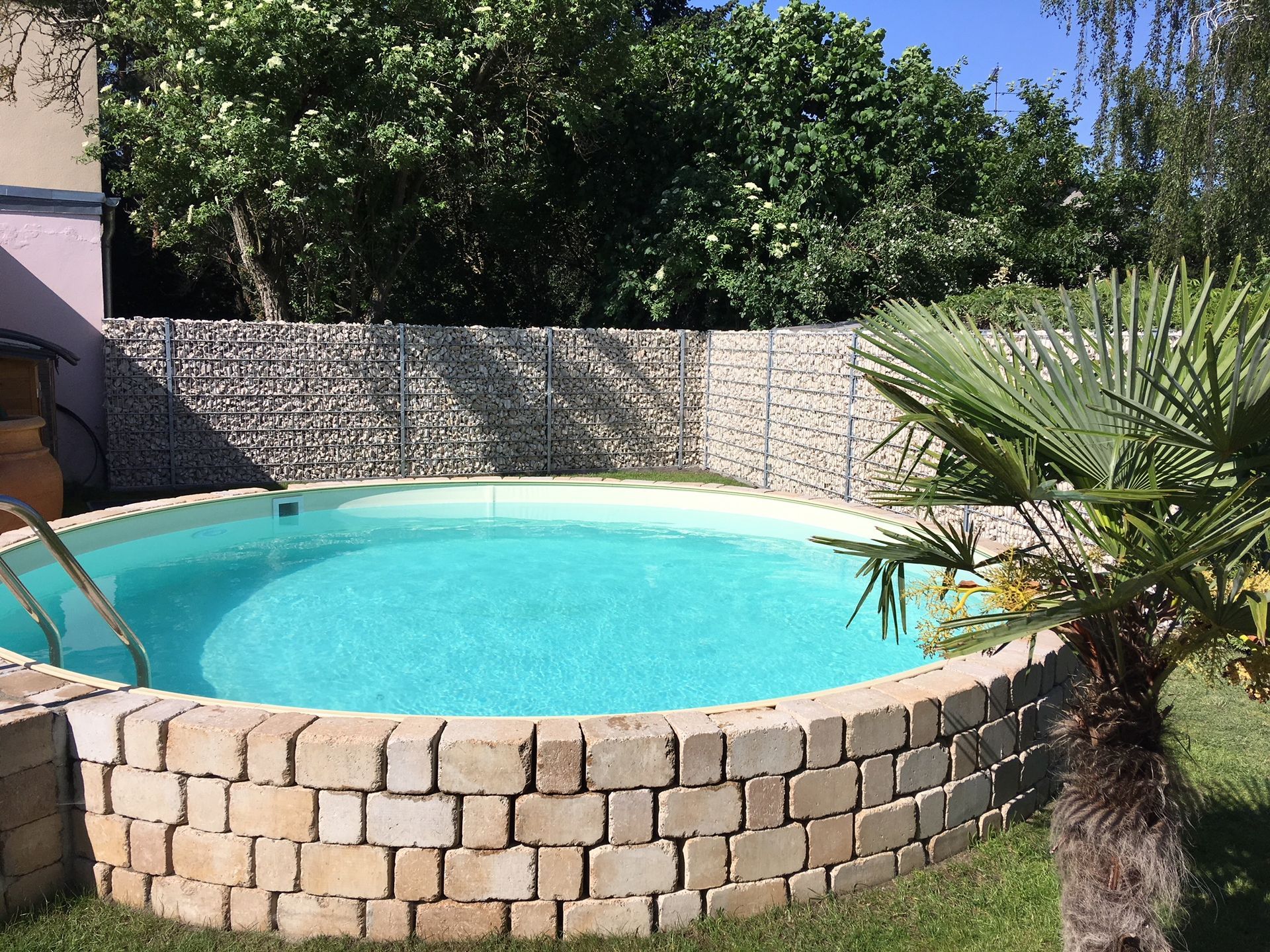 Ein runder, oberirdischer Pool mit türkisfarbenem Wasser, umgeben von Ziegeln und einer Felswand, in einem Hinterhof mit einer Palme und viel Grün.