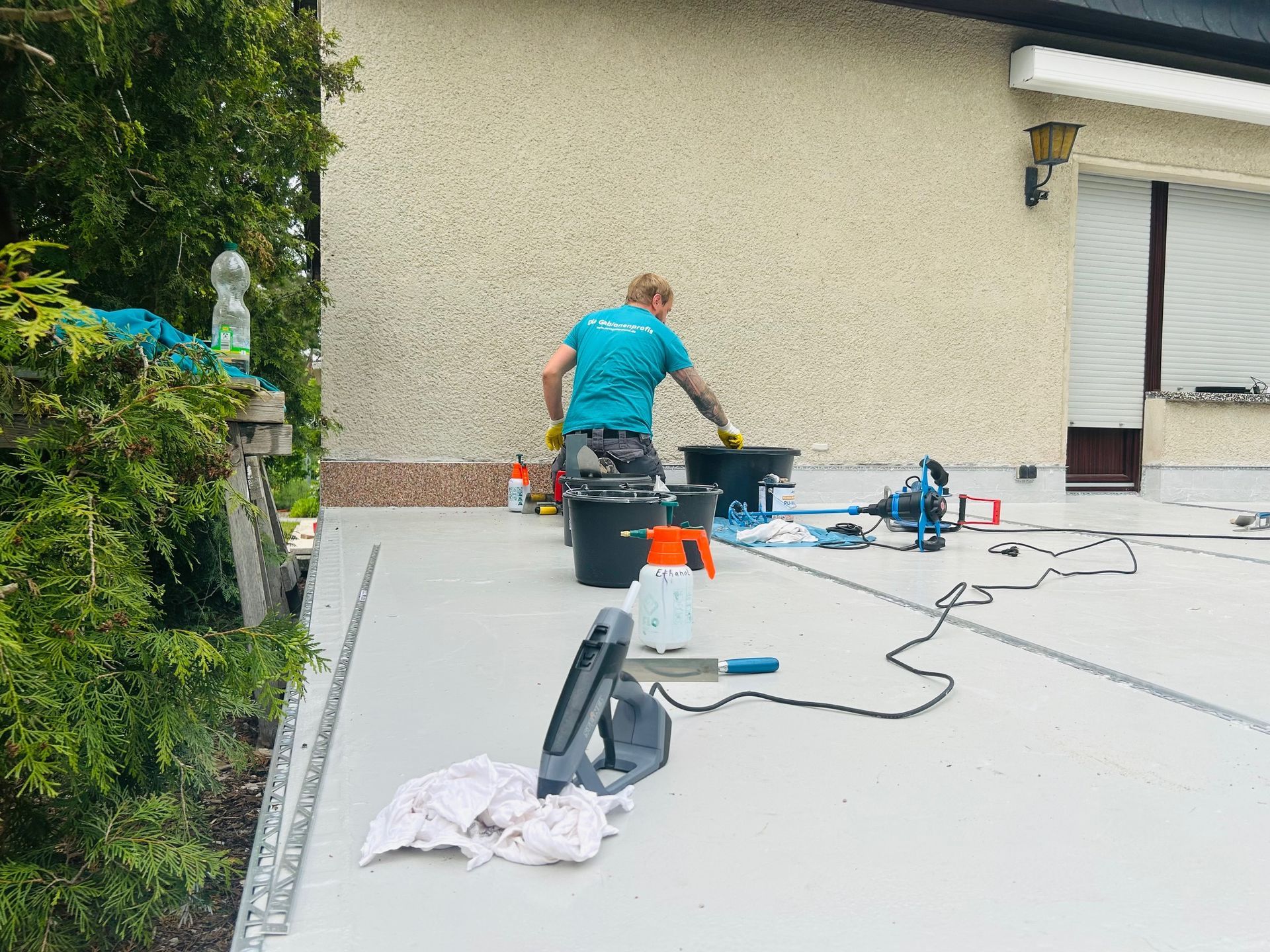 Eine Person rührt etwas in einem Eimer auf einem Flachdach. Verschiedene Werkzeuge, darunter eine Säge, liegen verstreut in der Nähe.