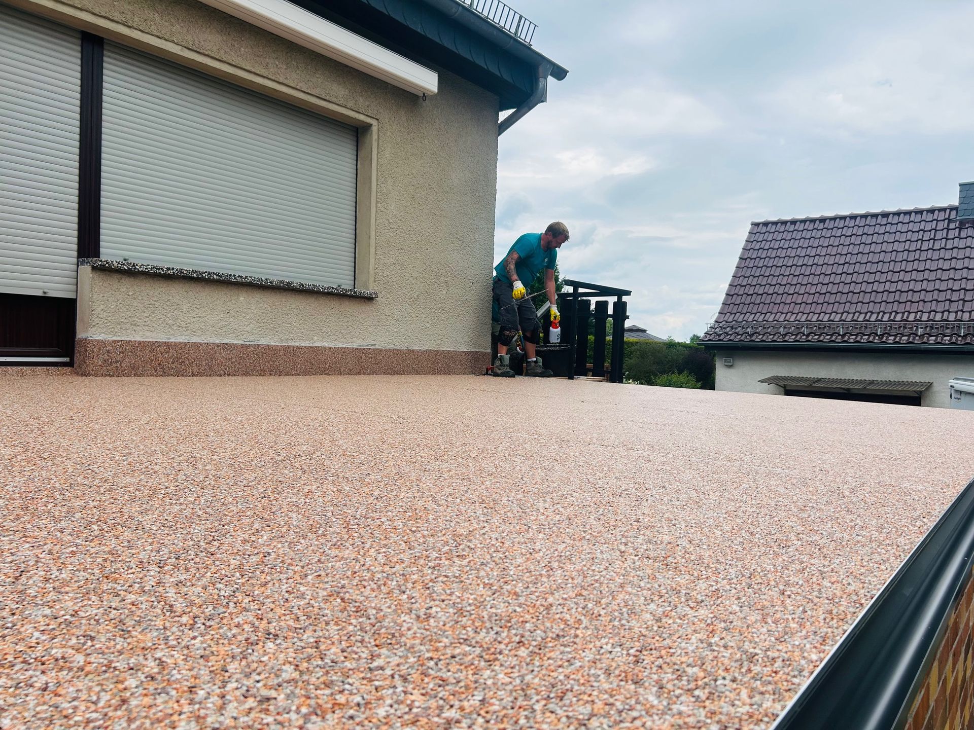 Person trägt Beschichtung auf ein mit Kies bedecktes Dach auf. Das Gebäude ist hellbeige, der Himmel ist bedeckt.