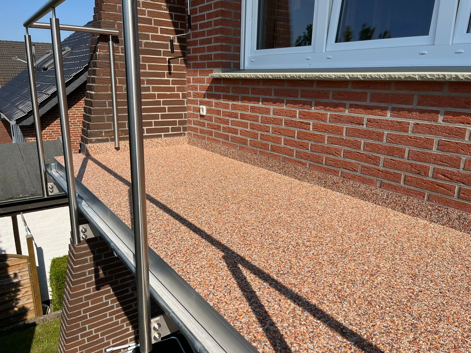 Eine mit rötlichen Kieselsteinen bedeckte Rampe führt zu einem Gebäudeeingang mit Edelstahlgeländer. Ziegelwände und ein Fenster sind sichtbar.