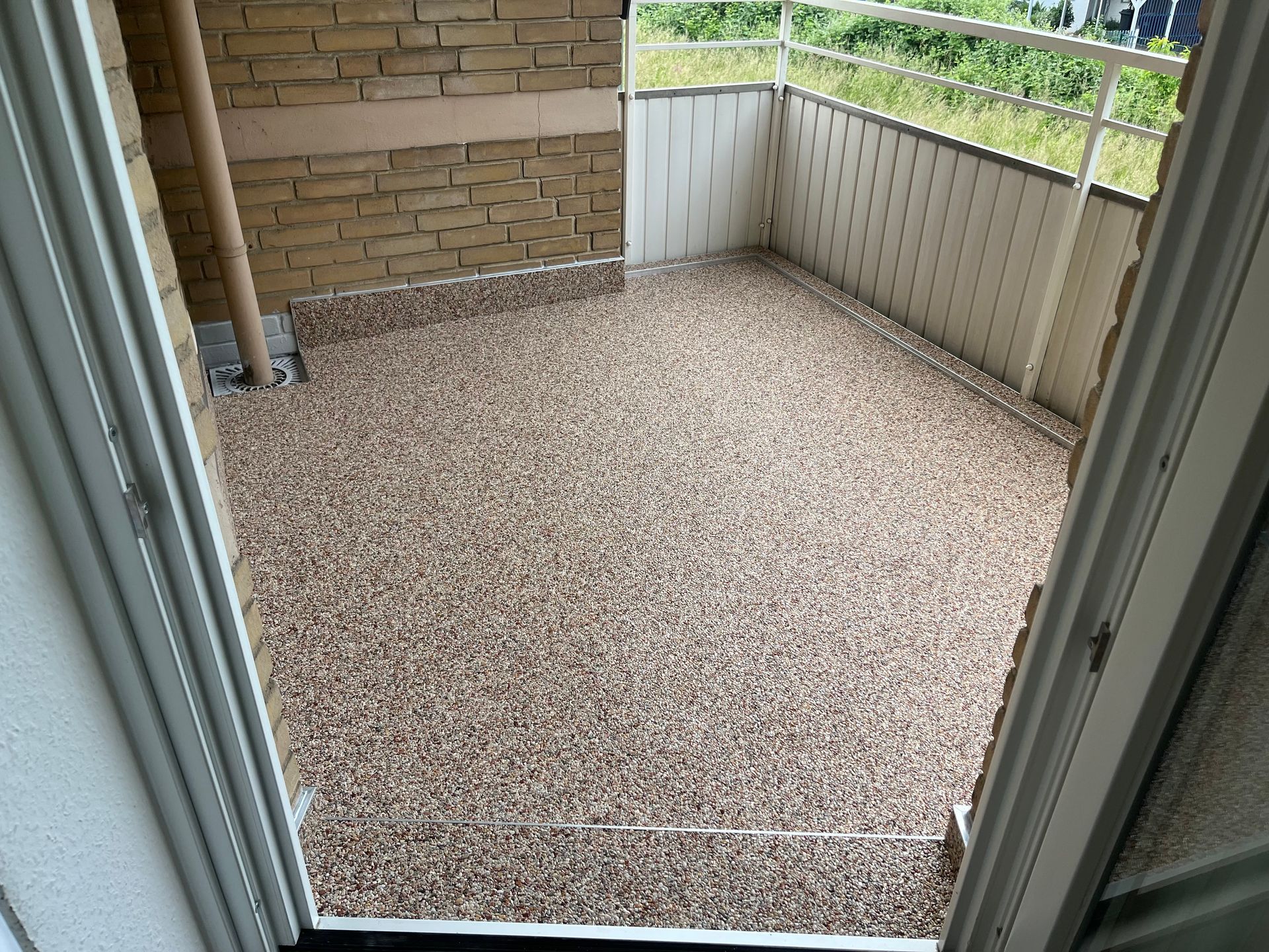 Kleiner Balkon mit kiesartigem Boden, beigen Wänden und weißem Geländer. Der Blick auf den Balkon erfolgt durch eine offene Tür.