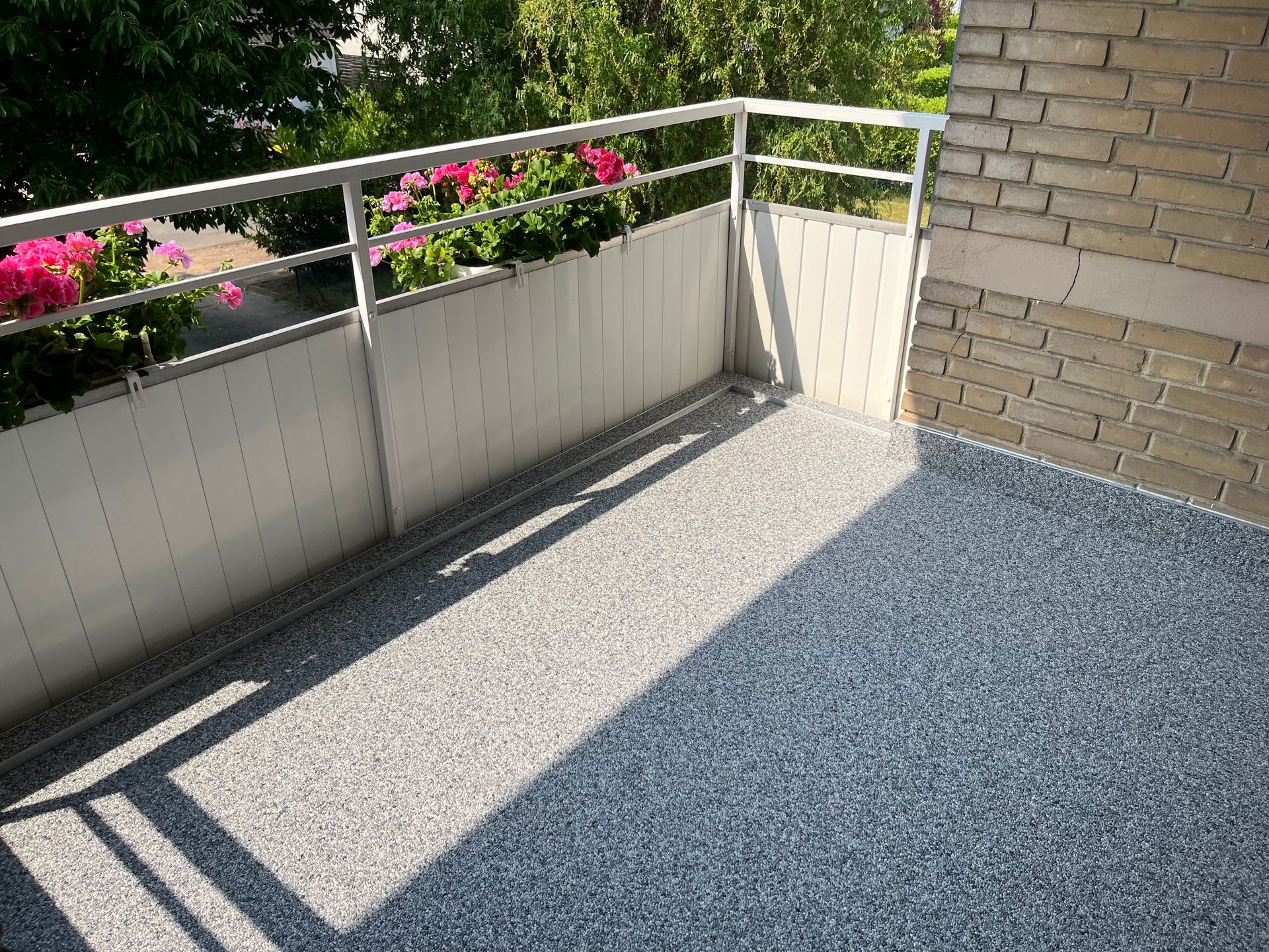 Balkon mit grauem Kiesboden, weißen Seitenteilen und einem Metallgeländer. Rosa Blumen im Hintergrund.